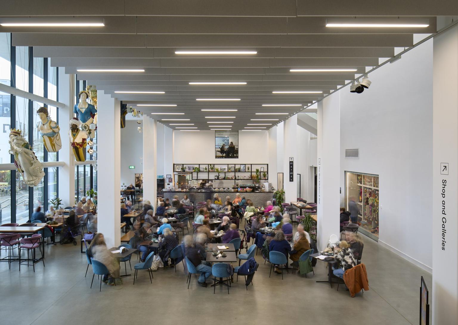 Busy modern café with blue chairs and high white ceilings where people sit and talk beneath hanging decorative sculptures and linear ceiling lights.