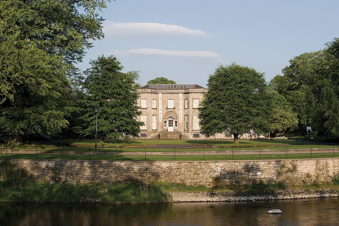 A grand house fronted by trees, seen from across a river