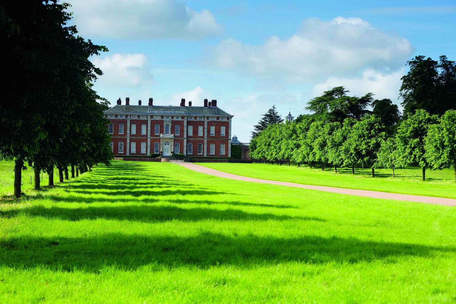 A long drive with lawns and a line of trees either side, leading to a stately red bricked house