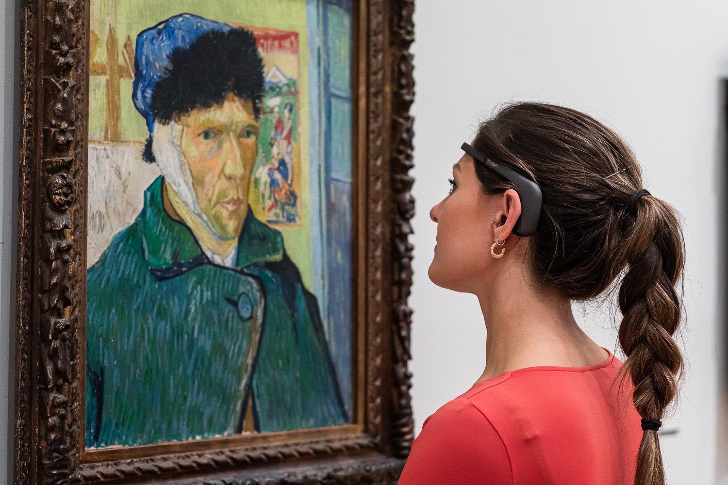 A woman wearing an EEG headset looks at a painted self-portrait by Van Gogh.