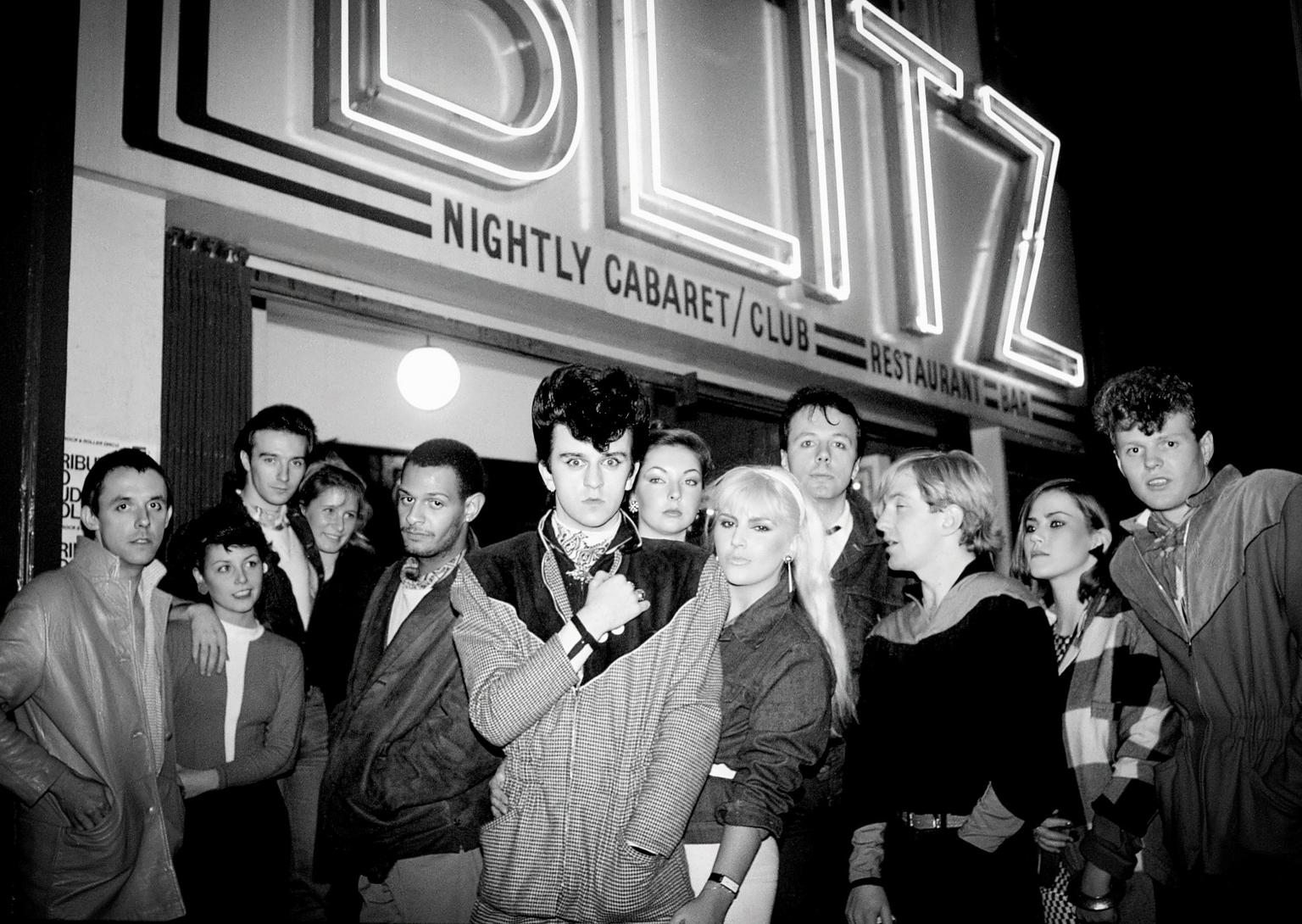 A group of young adults stand below a large neon sign saying 'BLITZ - nightly cabaret/club, restaurant, bar'.
