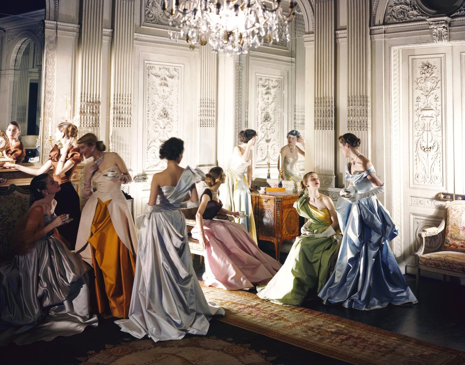 Women in elegant pastel ball gowns mingle while drinking tea under a crystal chandelier in a grand white room.