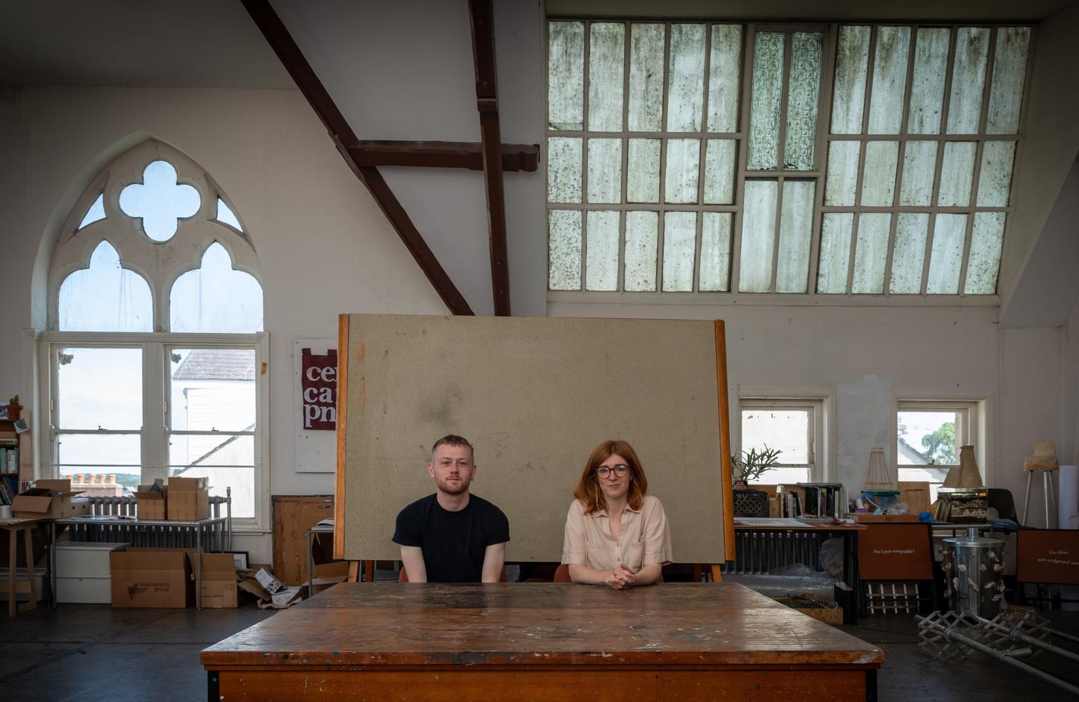 The artists Manon Awst and Dylan Huw sit behind a large wooden table in a bright, high-ceilinged studio with tall windows.