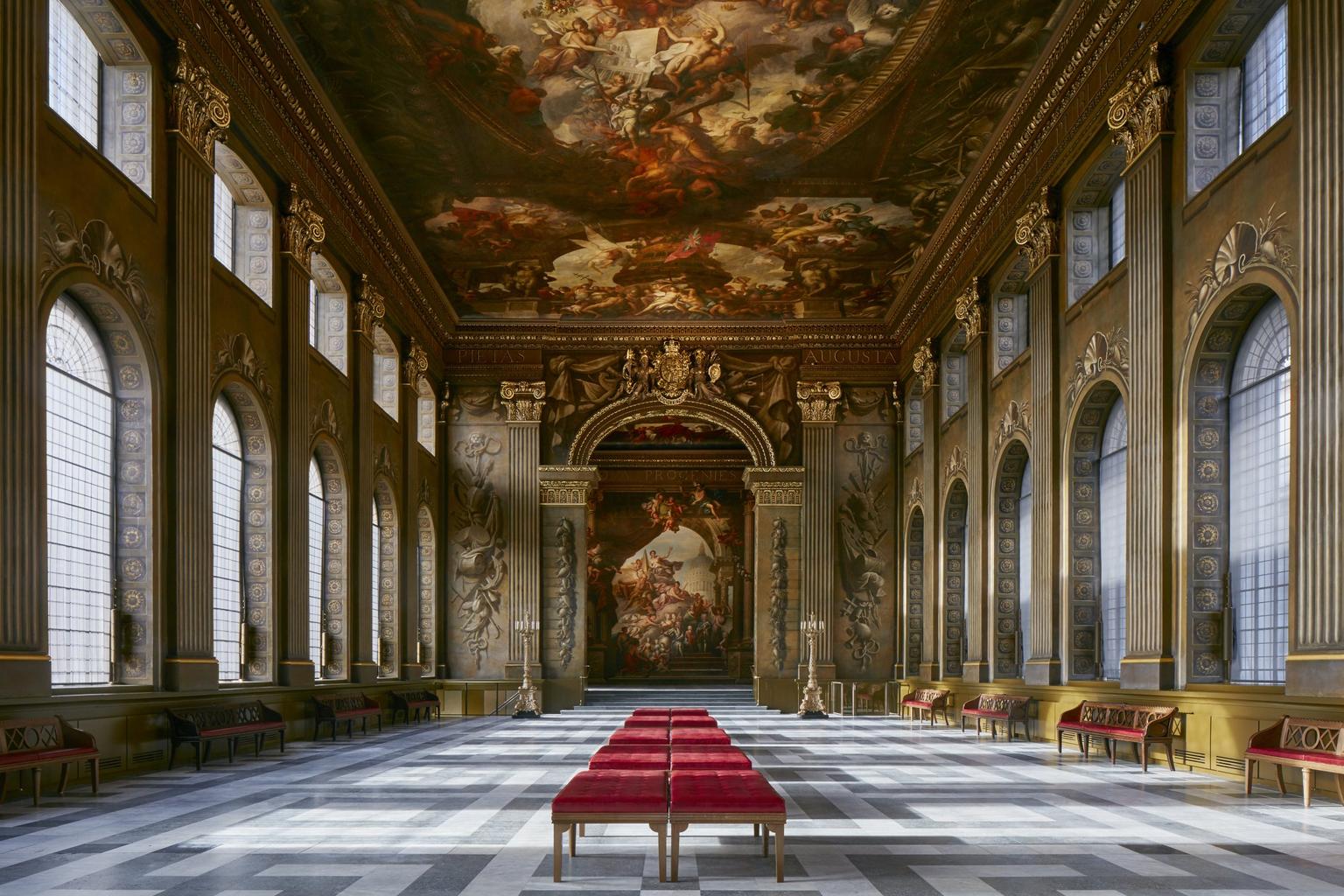 A grand hall, with painted ceiling, gold masonry, arched windows and red benches down the middle