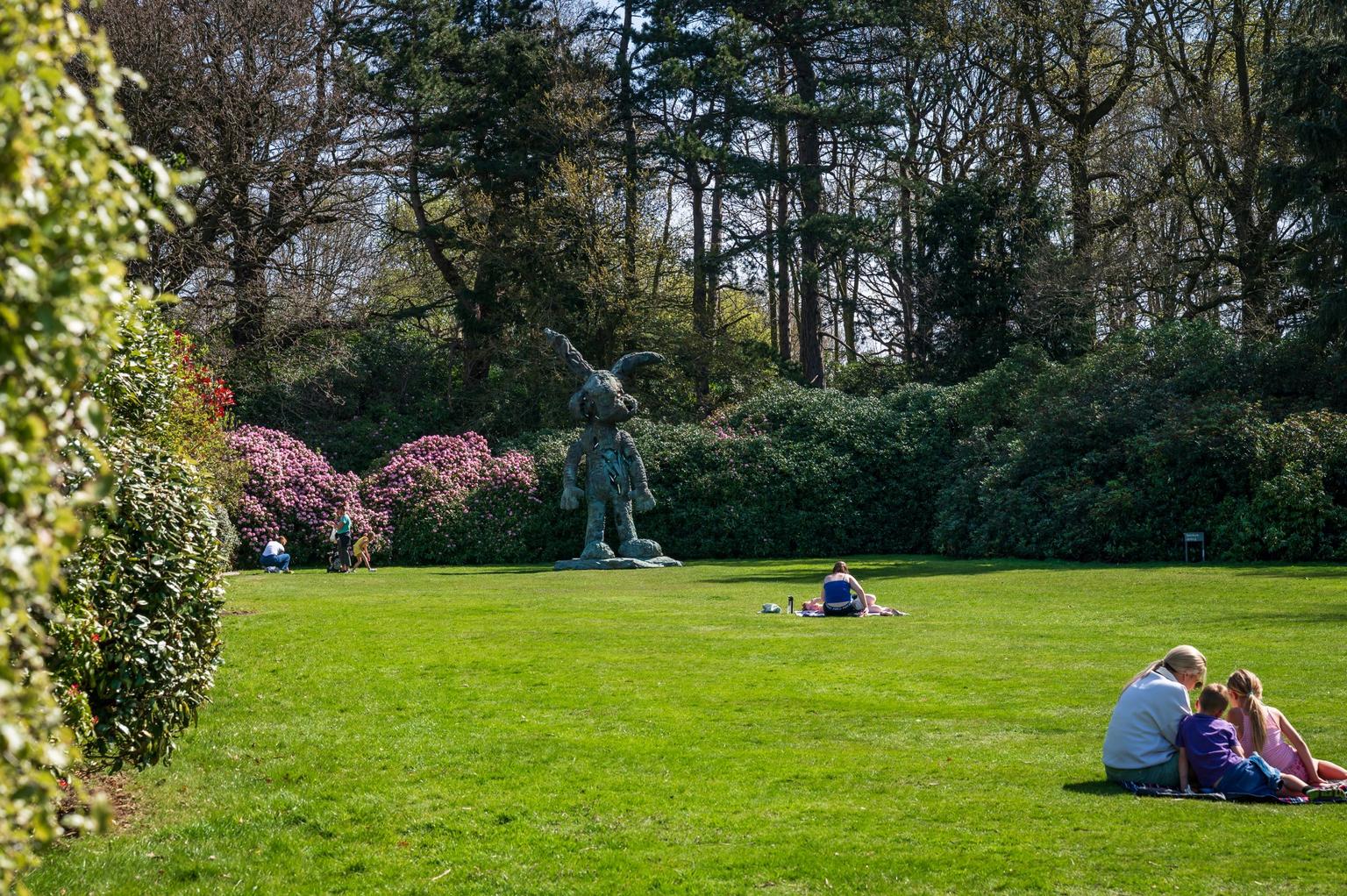 A metal bunny sculpture on a lawn surrounded by rhododendrons, with people sitting on it