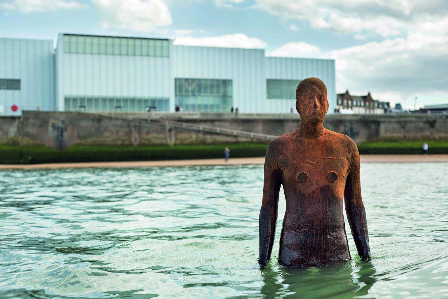 A rusted metal statue of a human figure up to its hips in the sea, with the Turner Contemporary in the background