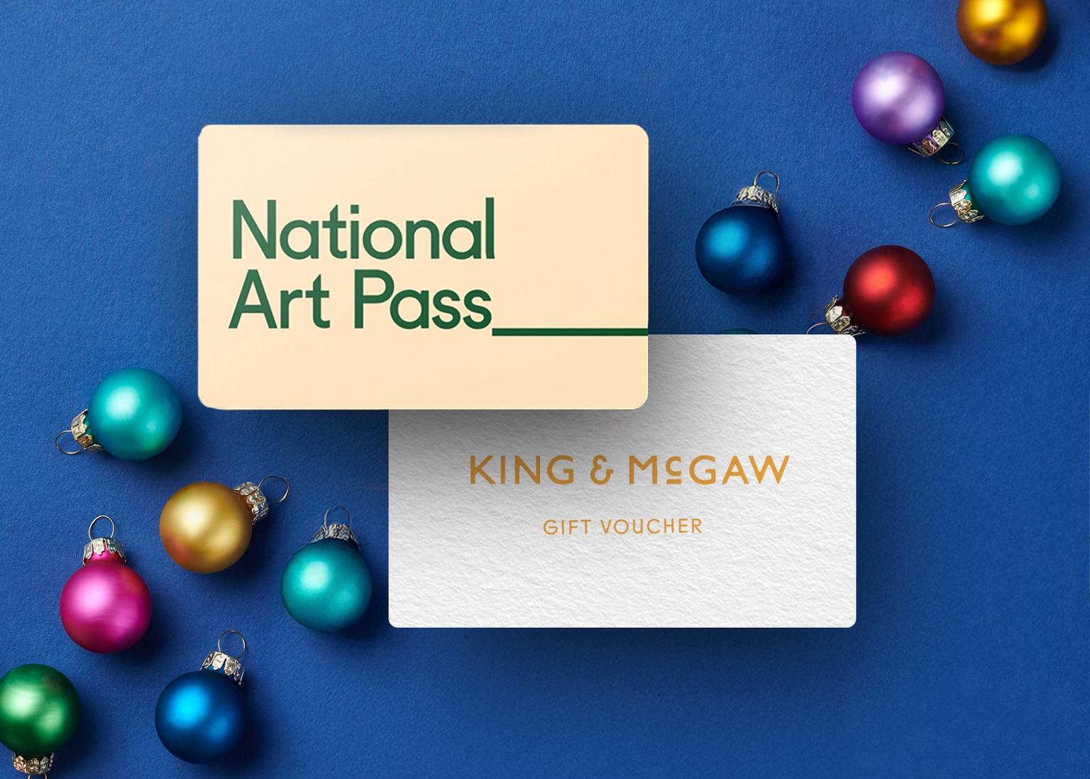 Two gift cards reading "National Art Pass" and "King & McGaw" placed on a blue background surrounded by colorful Christmas baubles.