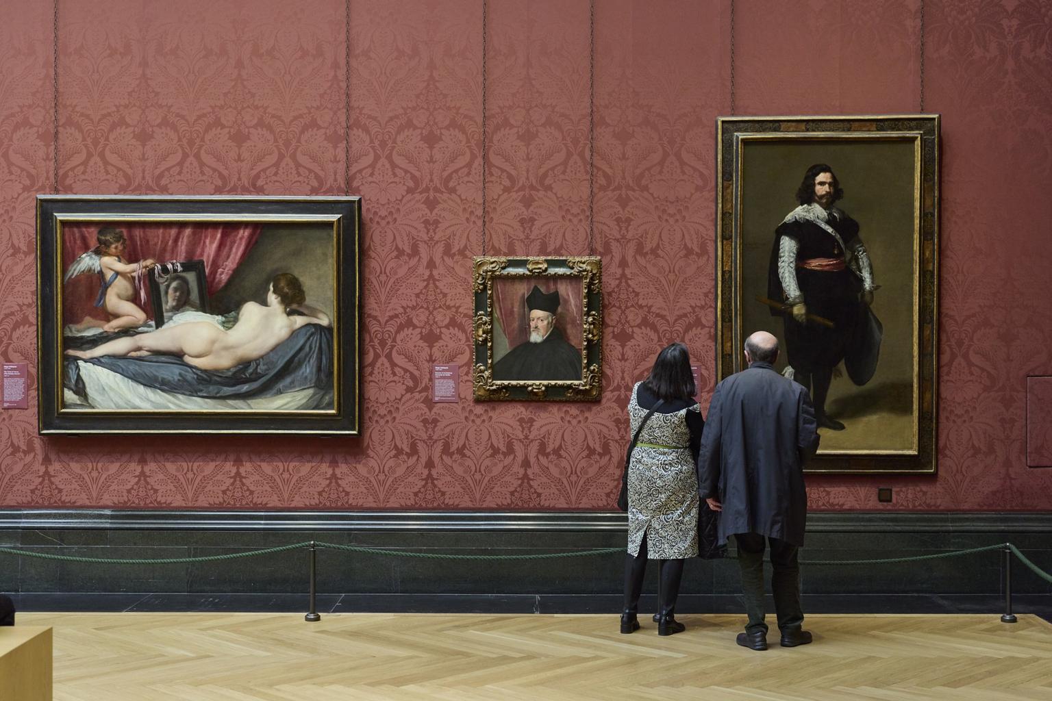 Two museum visitors stand before large framed paintings on a gallery wall decorated with red damask wallpaper.