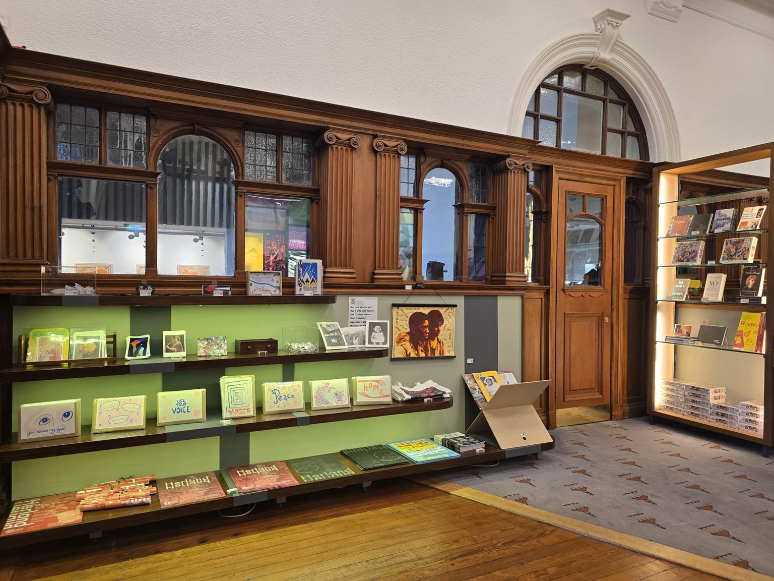 A museum gift shop counter with shelves displaying books, postcards, posters and prints against ornate dark wood panelling and an arched doorway.
