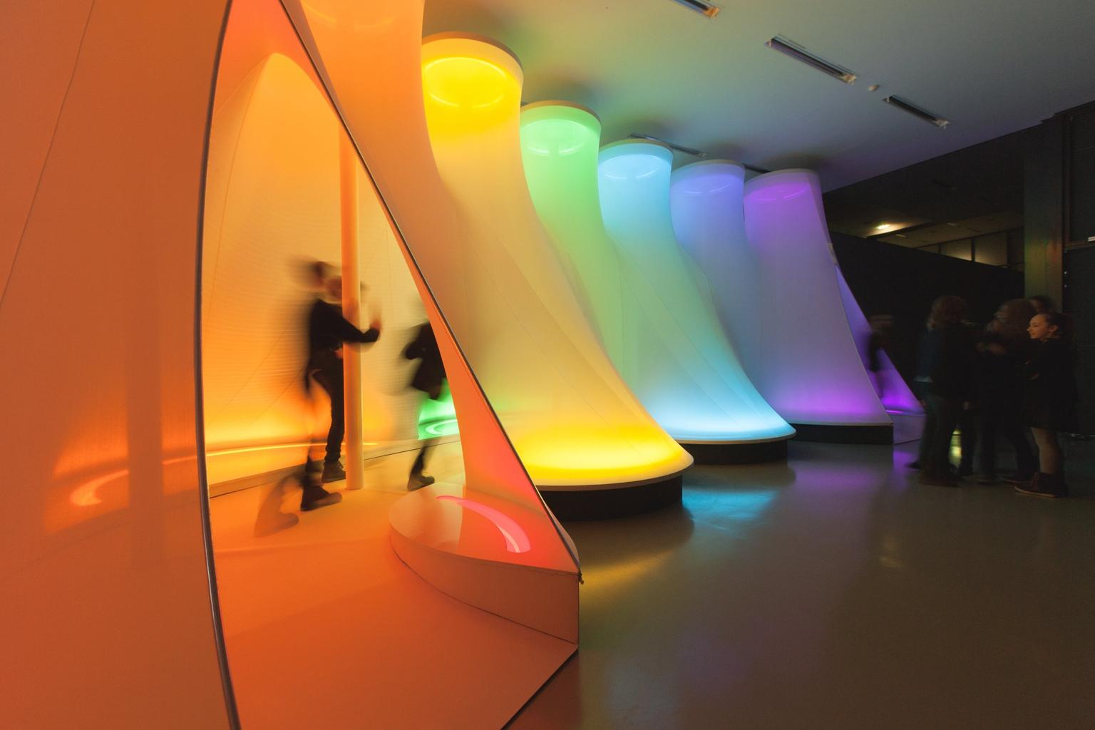 Visitors walk through a row of large glowing tubular art installations lit in rainbow colours in a dark exhibition space.