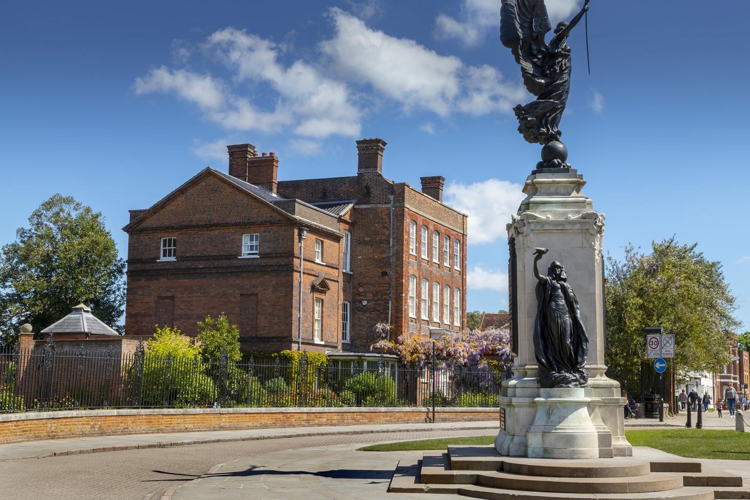 A red bricked, Georgian townhouse stands behind a metal fence; a neoclassical war memorial statute stands in a square in front of the house.