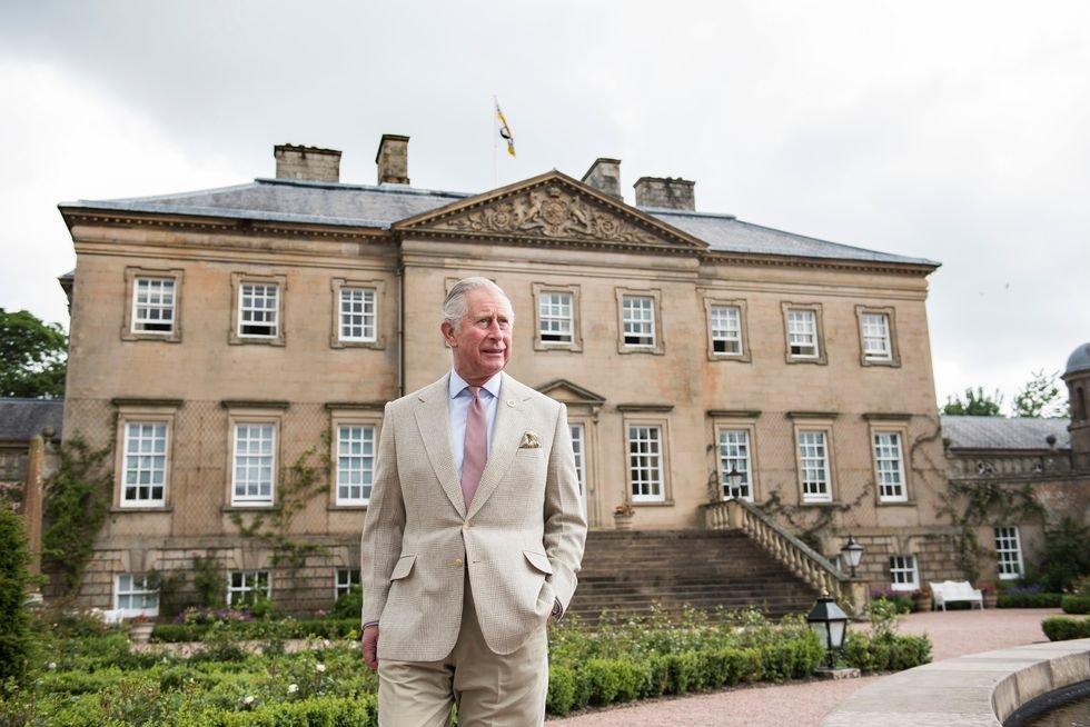 A picture of The King of England dressed in a beige suit, stood outside of a mansion