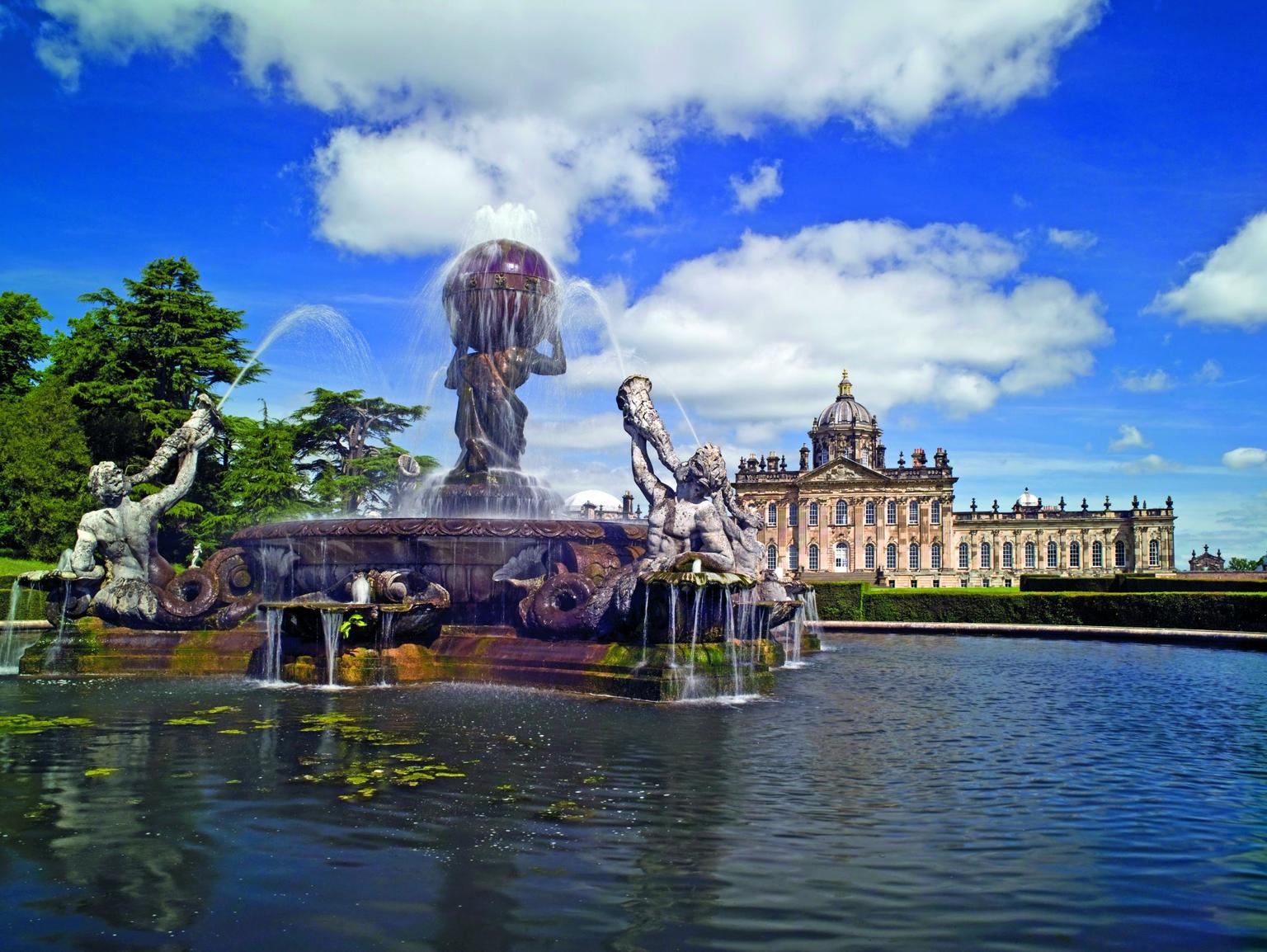 A water fountain of four male figures blowing water through horn-shaped shells, surrounding Atlas who holds up the globe. In the background is the grand historic house, Castle Howard.