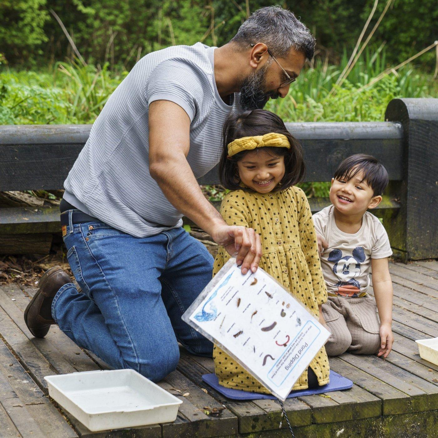 Two children and an adult crouch by the side of a pond, exploring the wild life that contained within it