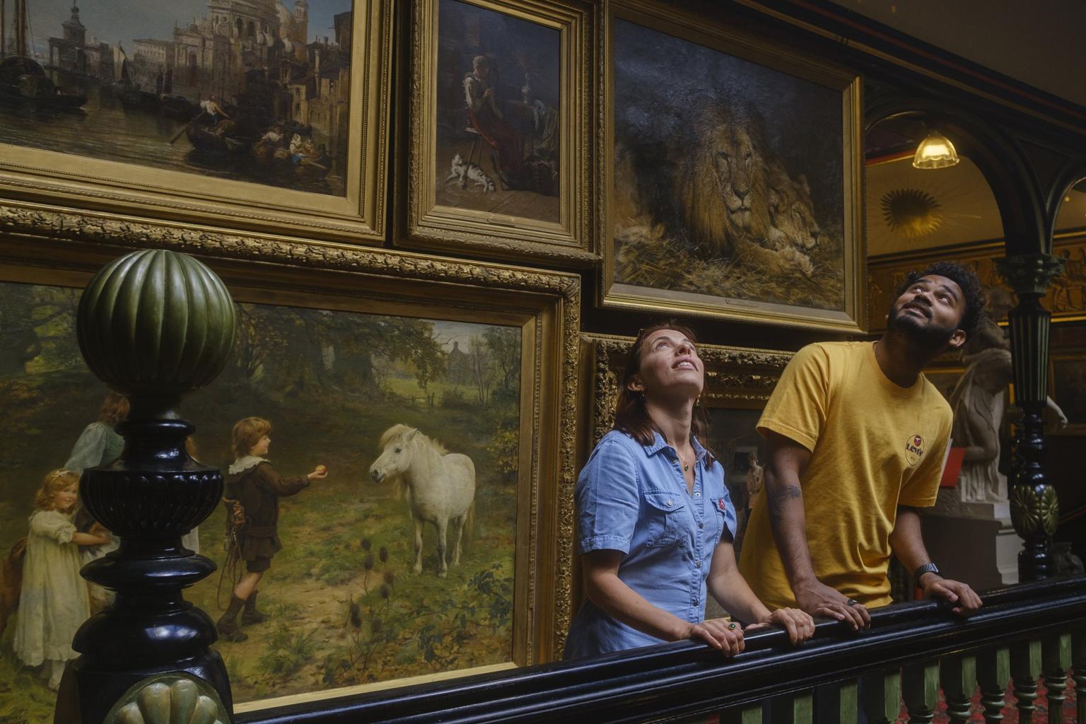 Two visitors lean against a banister and look up at the ceiling of a grand stairwell