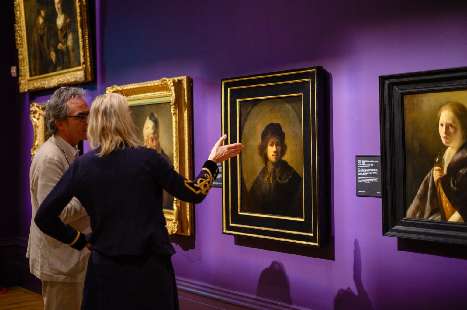 Two people discuss a framed portrait by Rembrandt on a purple gallery wall.