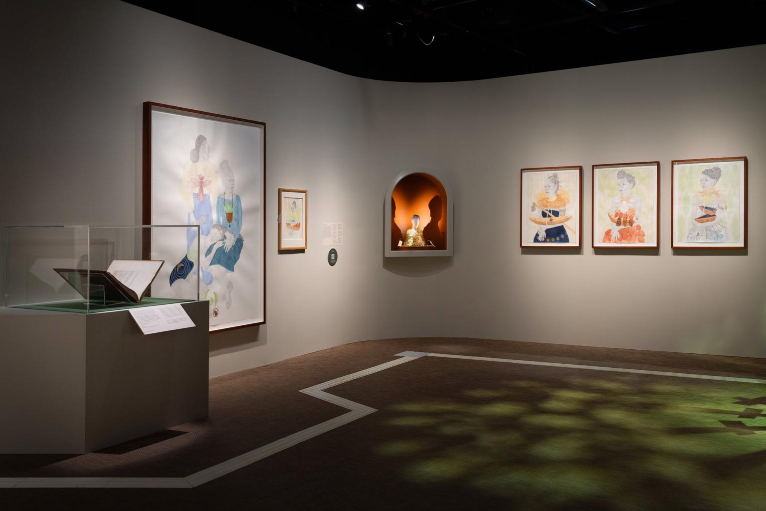 A dimly lit gallery displays framed portraits and an illuminated sculpture in an arched niche beside a glass case holding an open book.