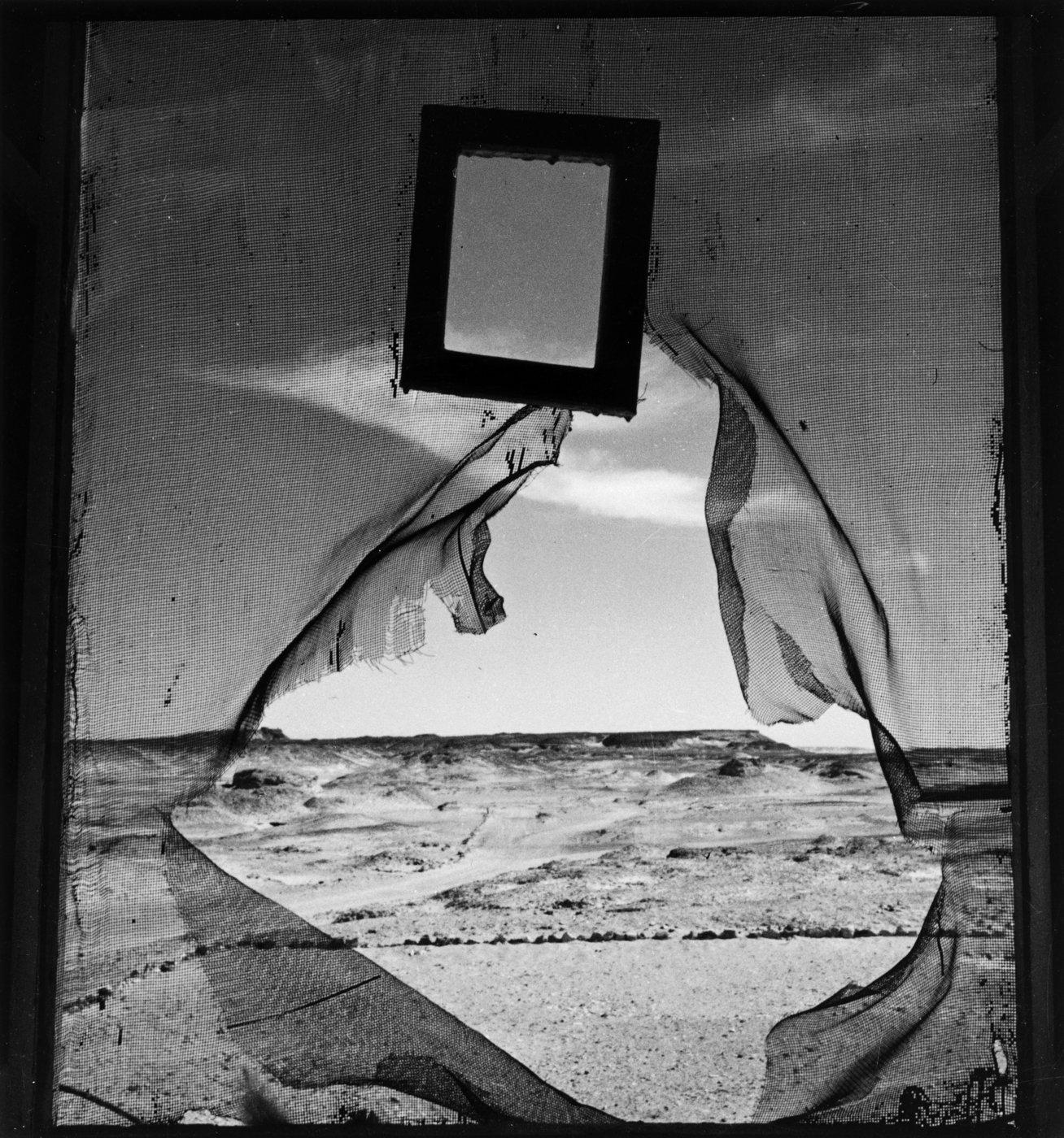 Torn sheer curtain billows around a broken window frame, revealing a windswept beach and choppy sea in stark black-and-white.