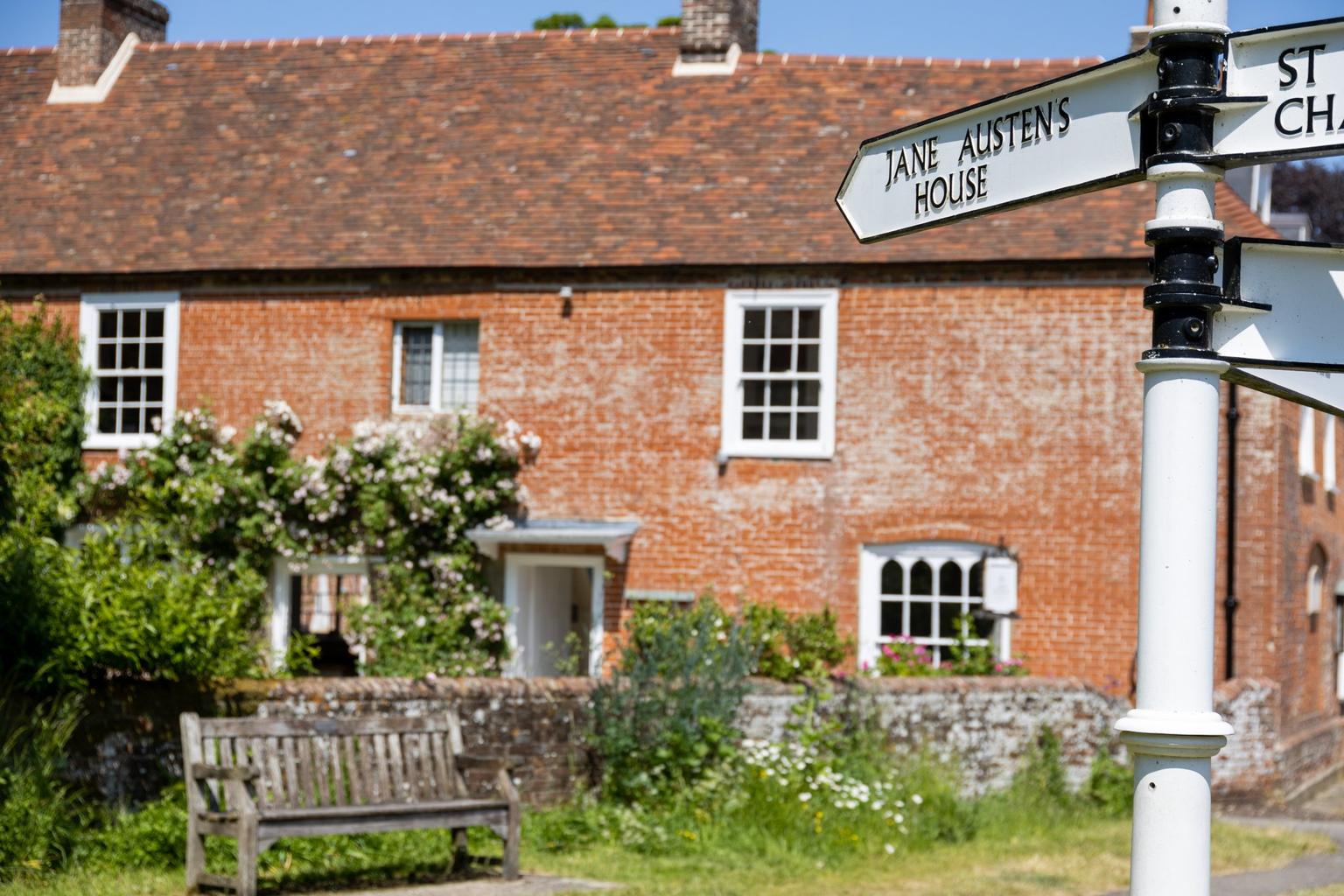 A black and white metal street sign pointing to Jane Austen's House, red brick with roses around the door