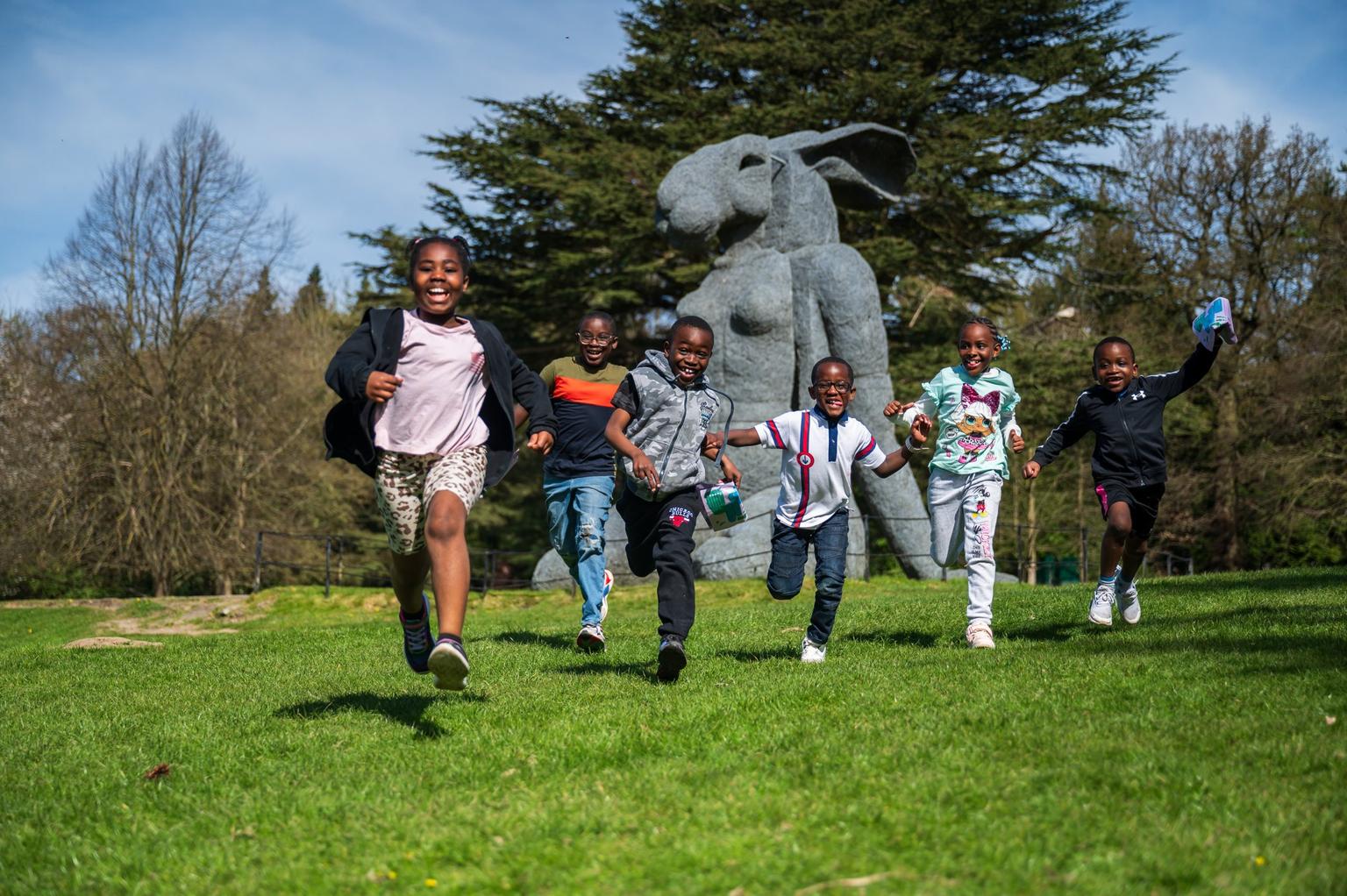 A group of children running joyfully on grass, in front of a large concrete sculpture of a rabbit.