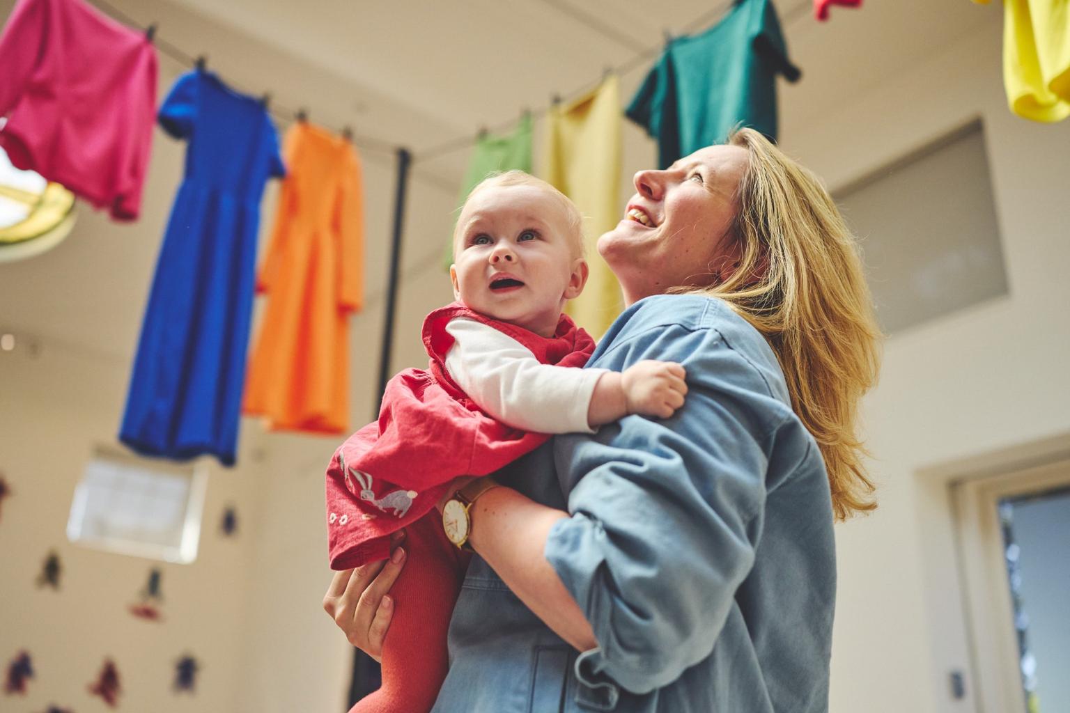 Smiling woman in a blue shirt holds a baby in a red dress beneath colorful hanging clothes.