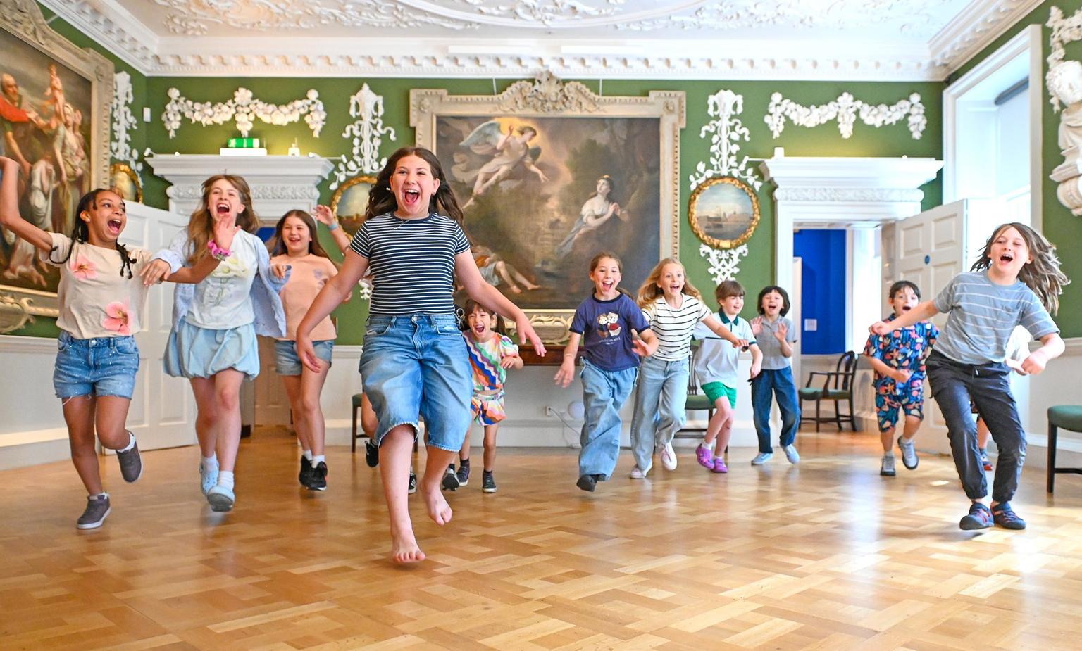 A group of excited children run toward the camera across a polished wood floor in an ornate green-walled gallery with large paintings.