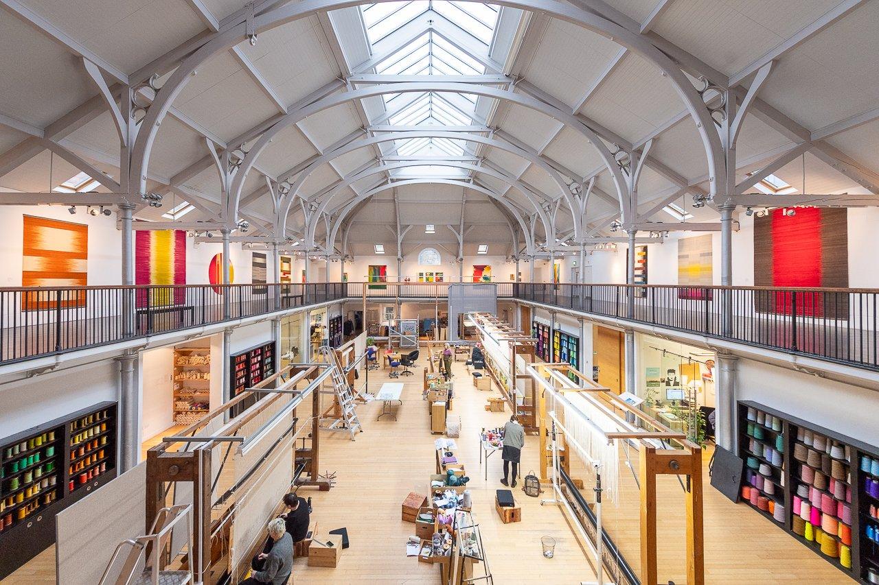 People working at looms in a weaving studios with tapestries on display at balcony level
