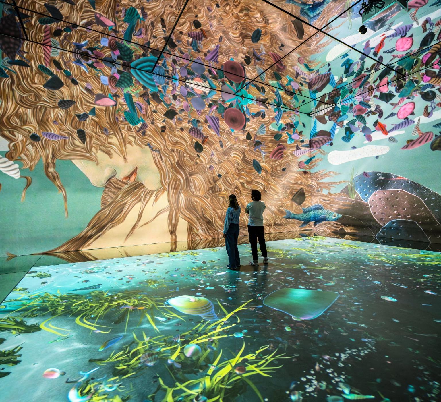 Two people stand in a turquoise immersive projection room filled with colorful floating fish and a giant flowing-haired face mural.