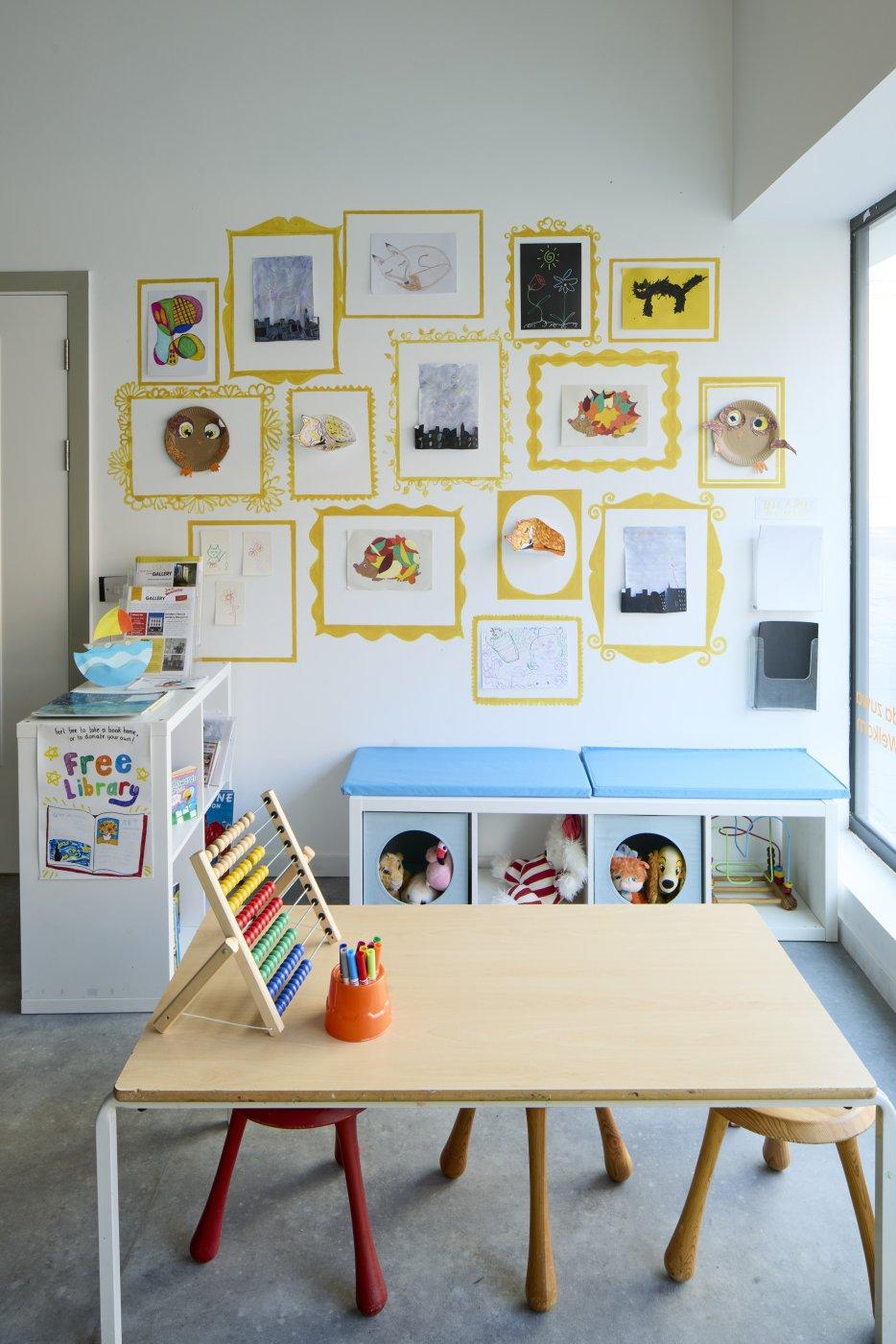 A children's play area with a small table and stools; an abacus and a pot of colouring pens are on the table. Children's artworks hang on the walls behind shelves of books, games and soft toys.