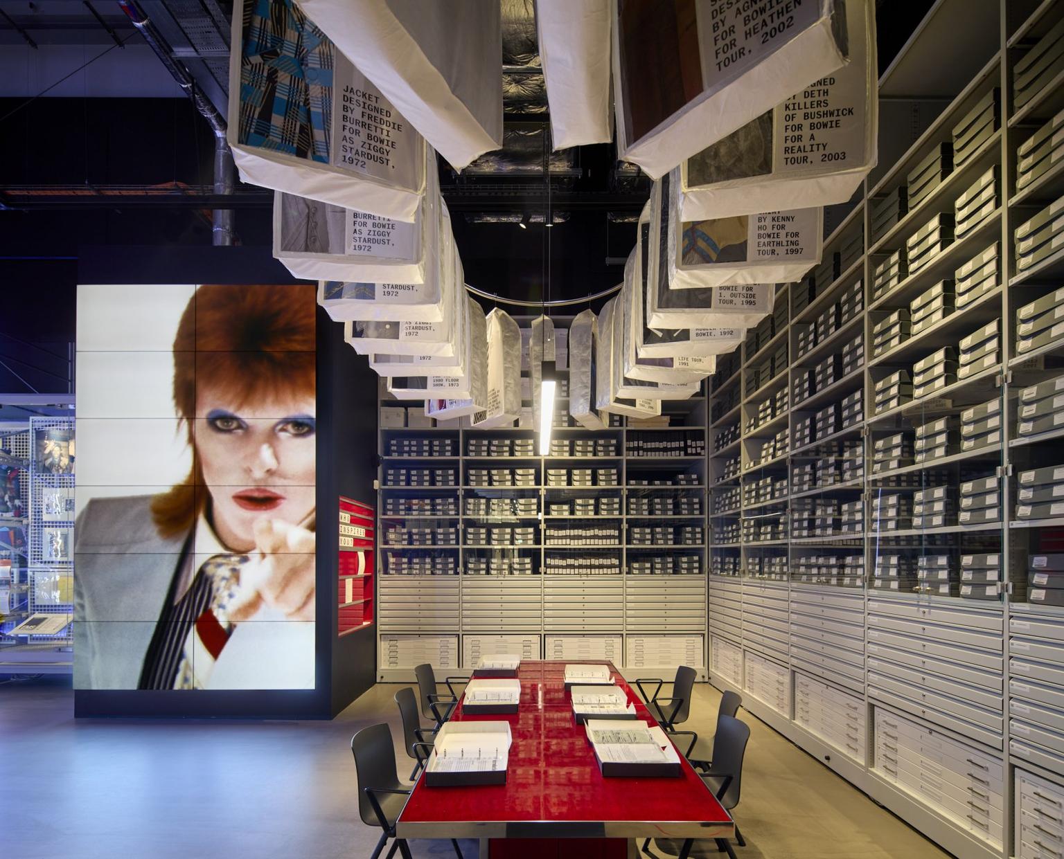 Interior archive room with a glossy red table and black chairs under hanging white banners, surrounded by gray filing shelves and a large images of David Bowie on a digital screen.