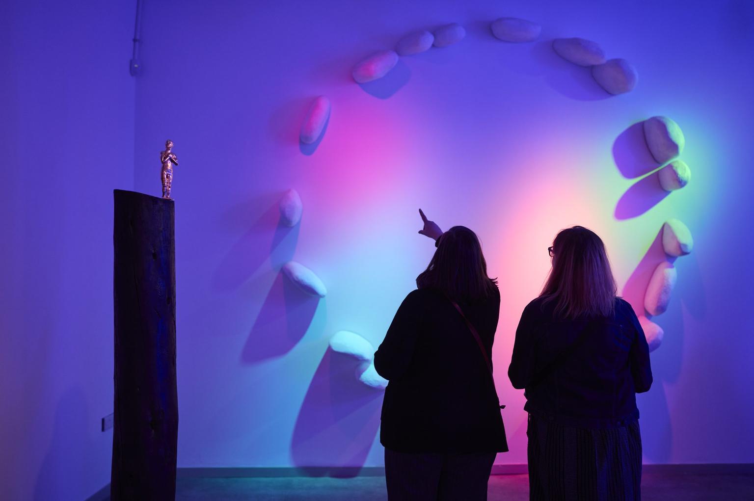 Two women stand in a dimly lit gallery space, looking at a sculpture of stones in a circle protruding from the wall, and a miniature gold figure on top of a black plinth. Pink, blue, green and orange lights project onto the white wall.