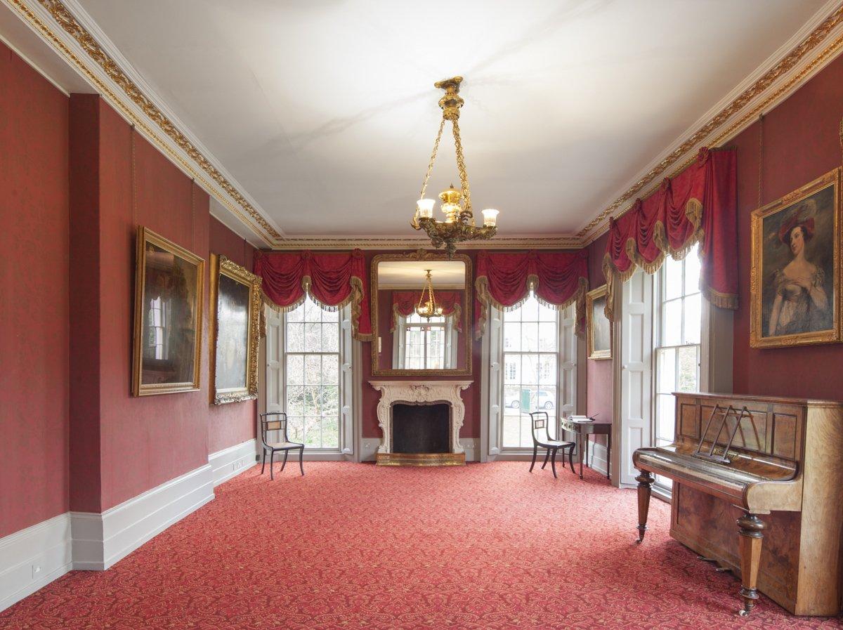 A Regency drawing room with red walls, curtains and carpet, featuring a fireplace and piano
