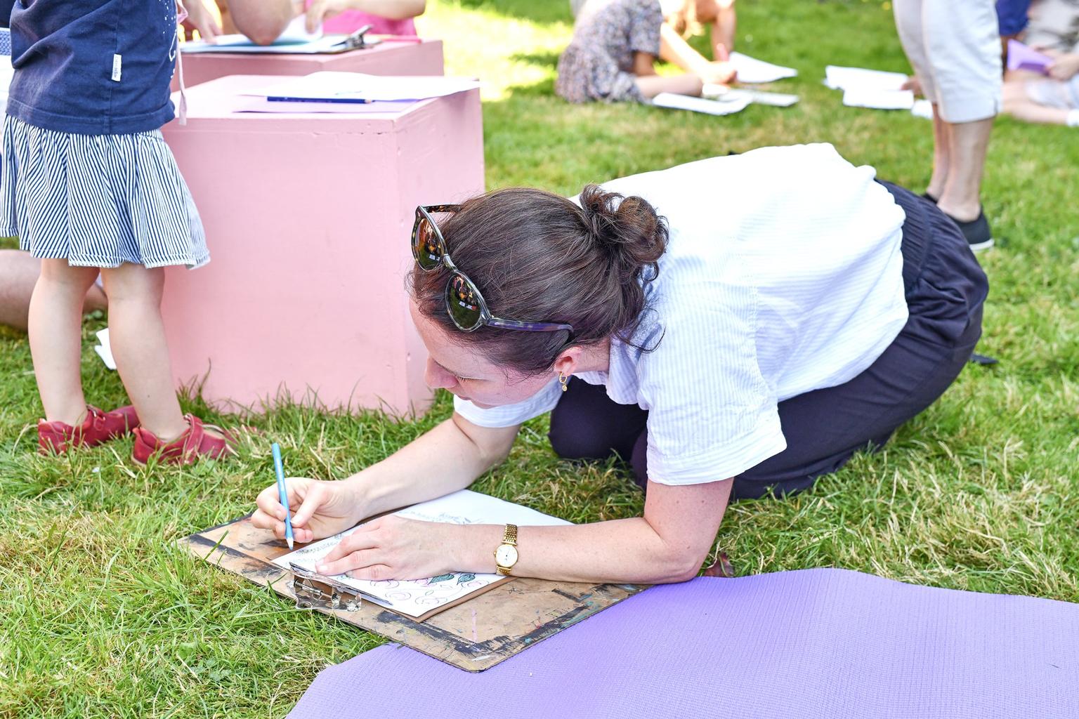 A woman kneels on grass to draw on paper attached to a clipboard with children playing in the background