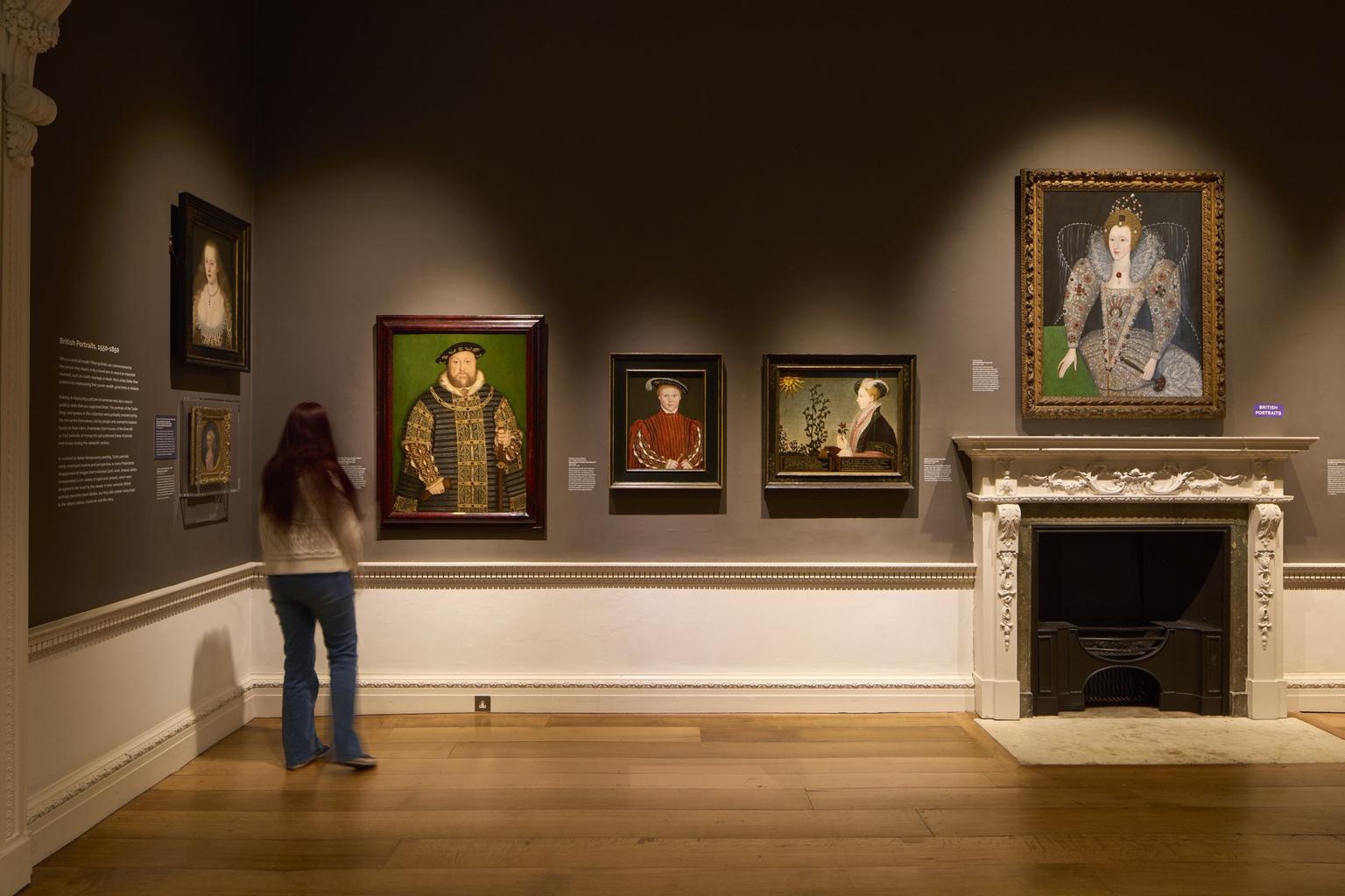 A woman looks at portraits of Henry VIII, Elizabeth I and King Edward VI hanging on a brown walled gallery space with an ornate fireplace.
