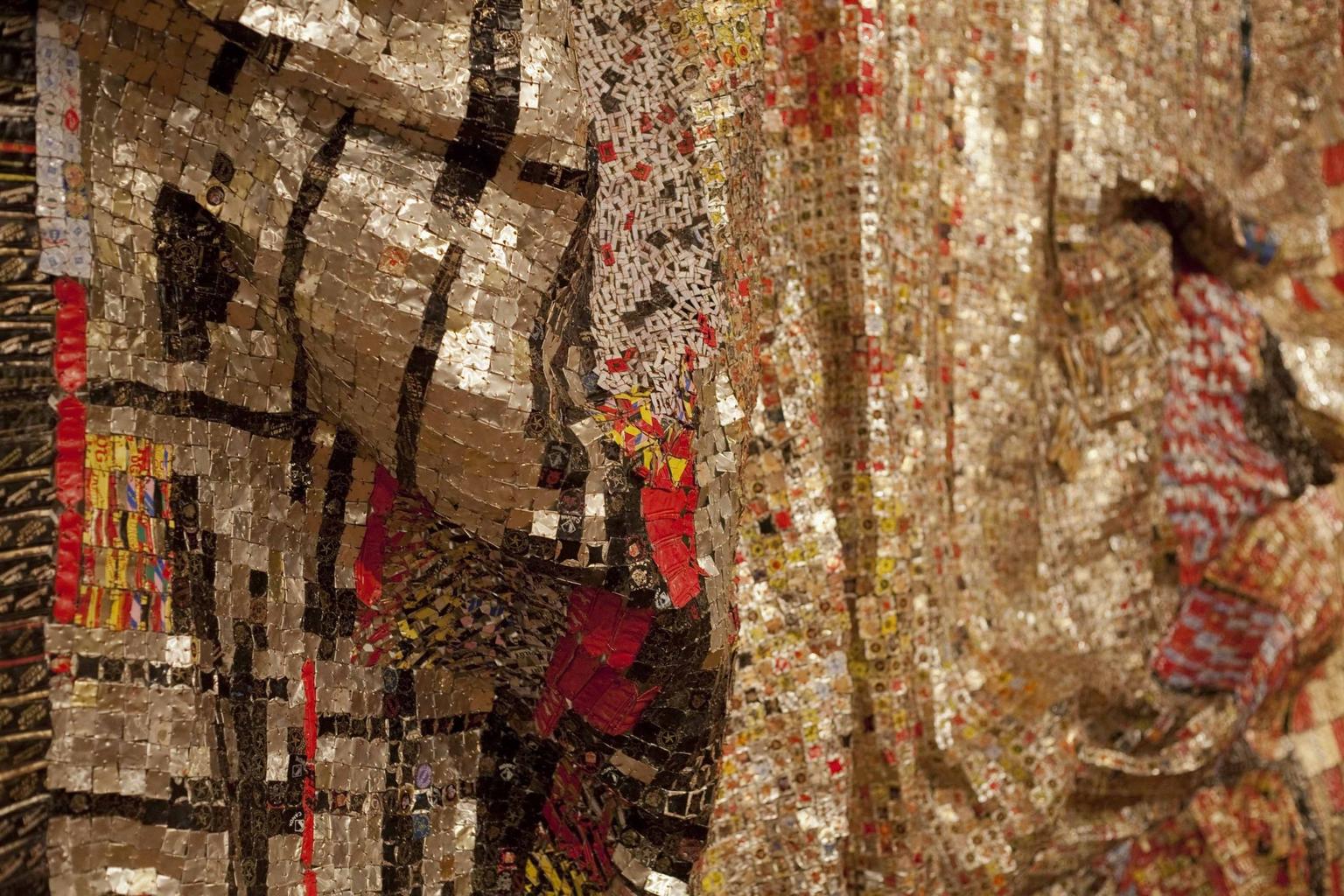 A close-up view of a large hanging work made with small pieces of mostly gold-, black- and red-coloured aluminium, held together with copper wire.
