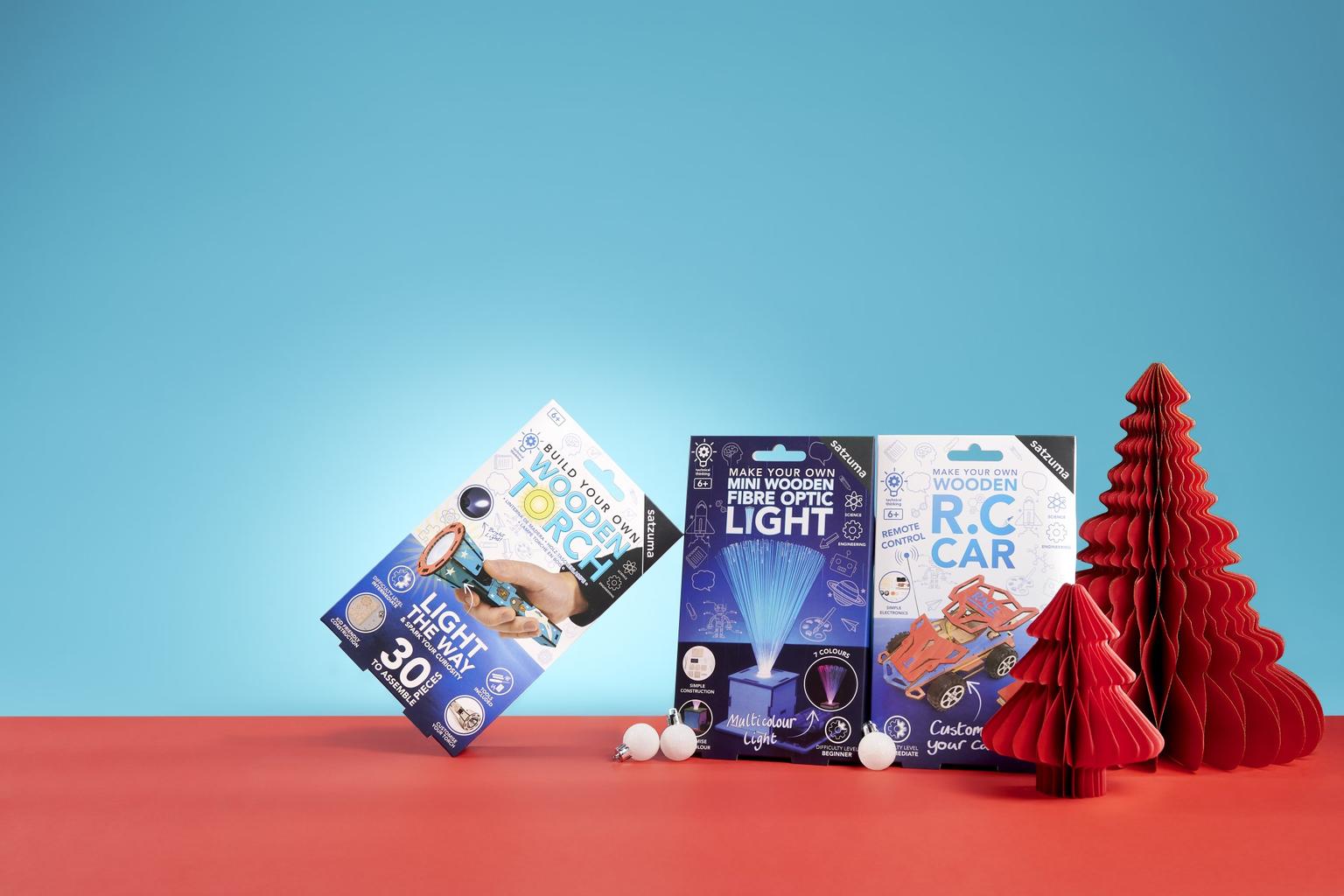 Three DIY science kits and red paper Christmas trees arranged on a red surface against a cyan background.