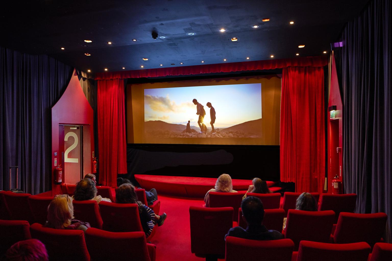 A group of people sitting in a cinema watching a film. There is a red carpet on the floor, matching red seats and red curtains frame the screen.