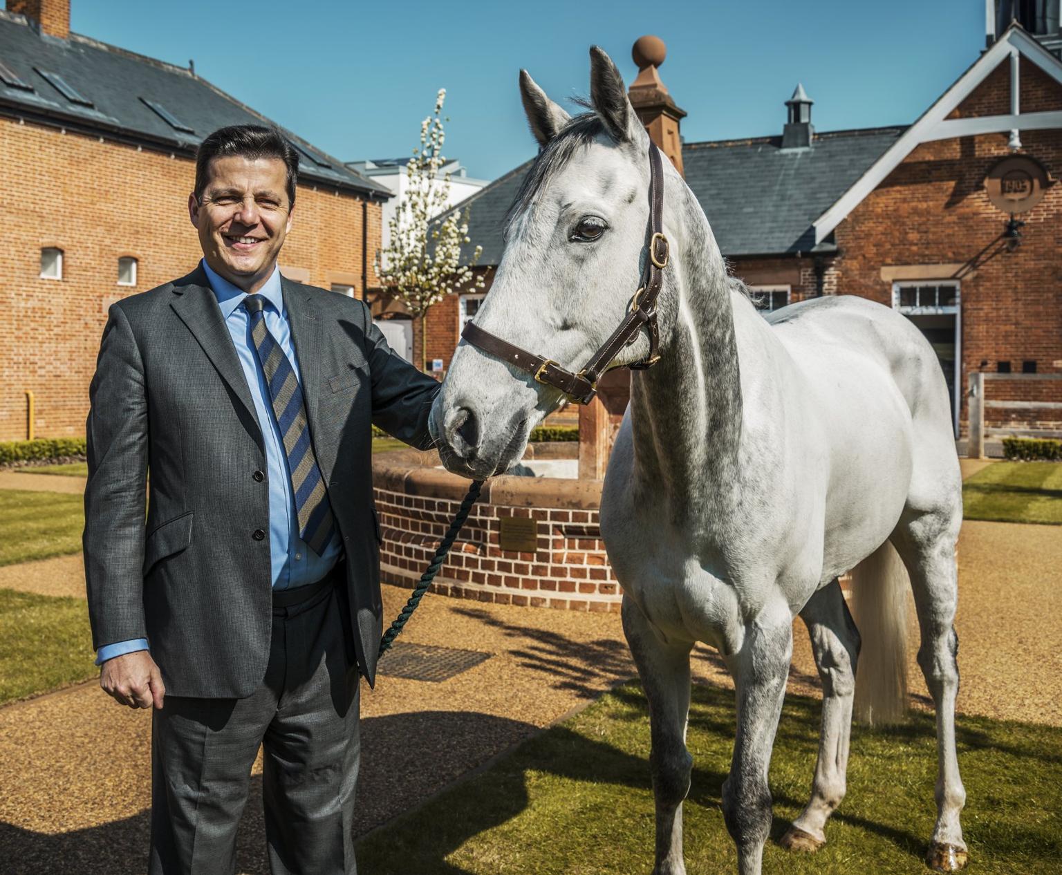 A man in a suit holding the lead rein of a grey horse.