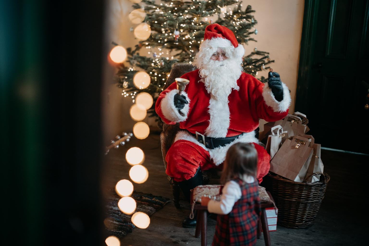 Santa Claus rings a bell while sitting by a decorated Christmas tree as a young child watches.