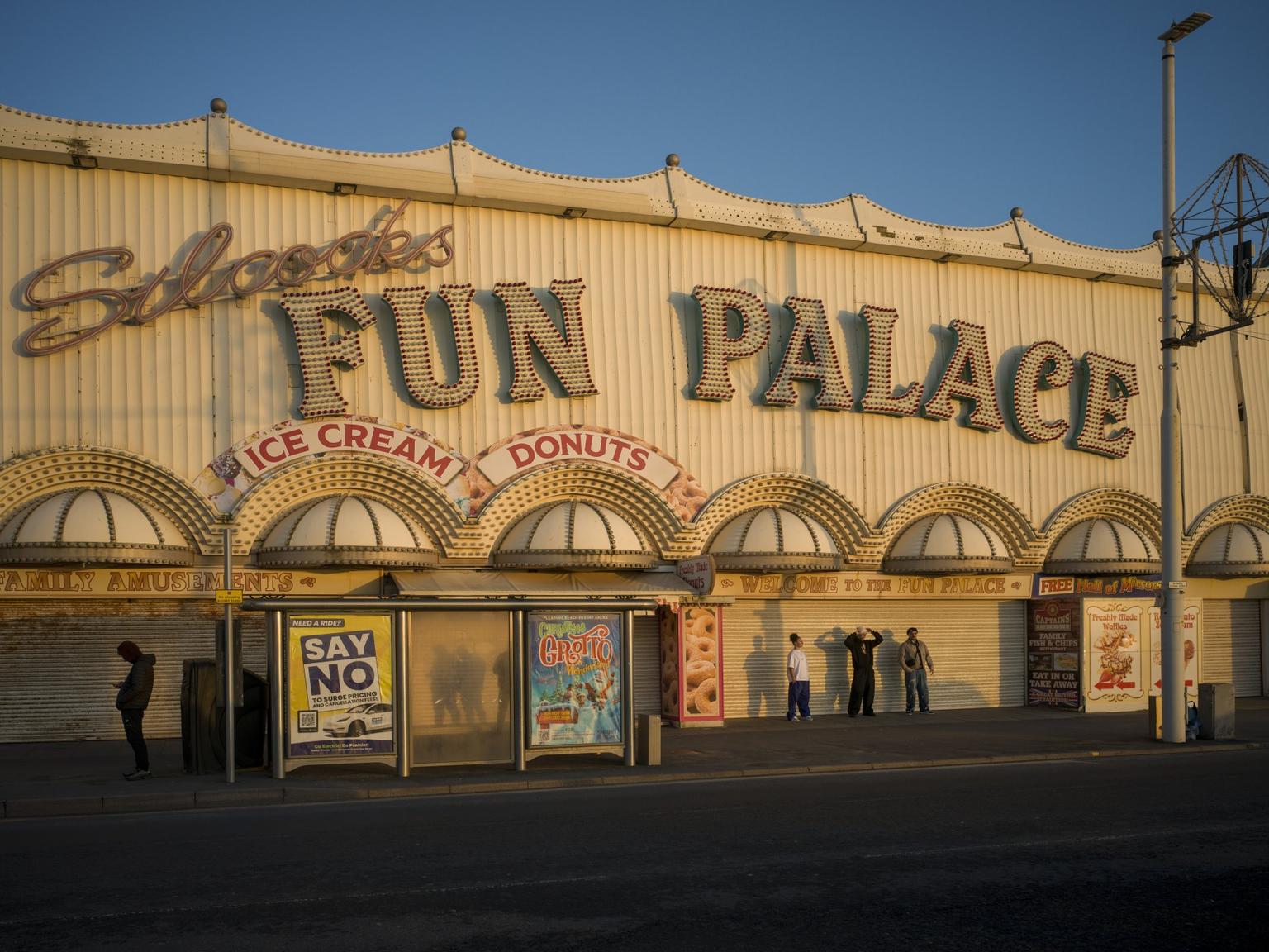 The facade of Siclock's Fun Palace, an amusement arcarde in Balckpool, bathed in golden light with a bus shelter and a group of teenagers in front.