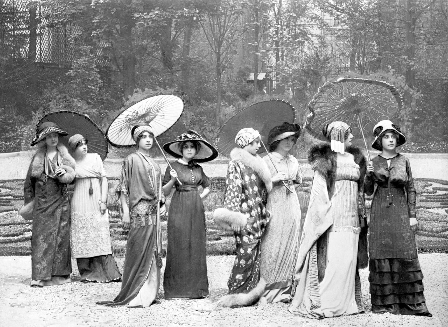 Black and white photograph of a line of elegantly dressed women posing with parasols in a formal garden.
