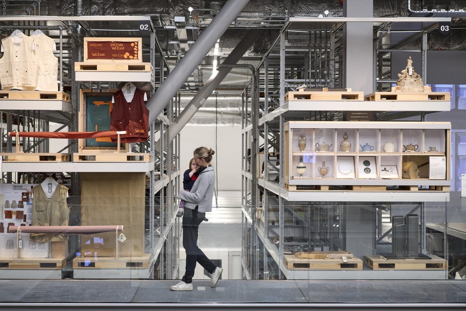 A woman carries a child while walking along a glass walkway past rows of museum storage shelves filled with artifacts.