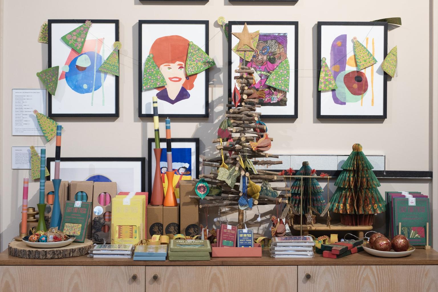 A shop display of colorful handmade holiday decorations, including a twig Christmas tree and patterned paper trees in front of bright framed artwork.