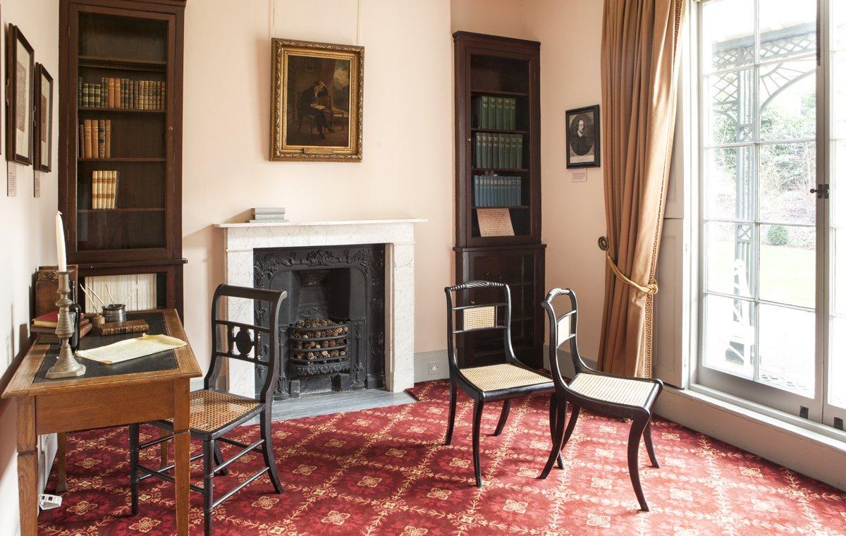 A drawing room with antique desk, bookshelves, fireplace and red patterned carpet.