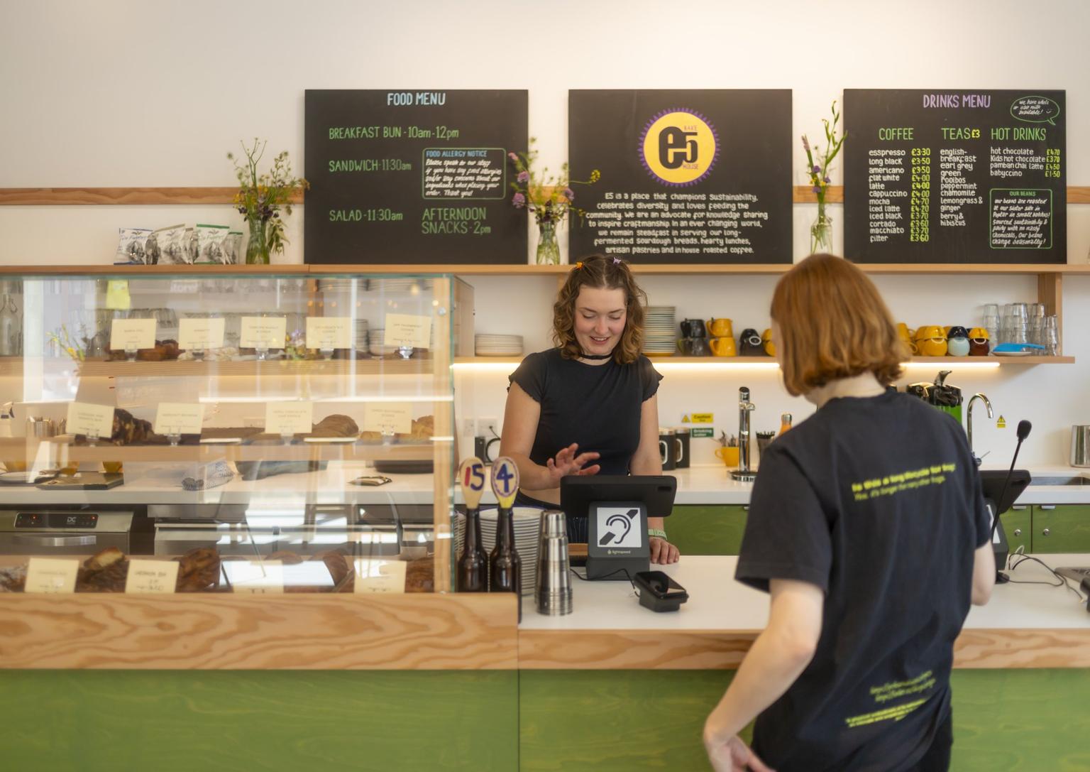 Barista in a black shirt taps a tablet at a bright café counter with green paneling and a glass display case filled with pastries while serving a customer.