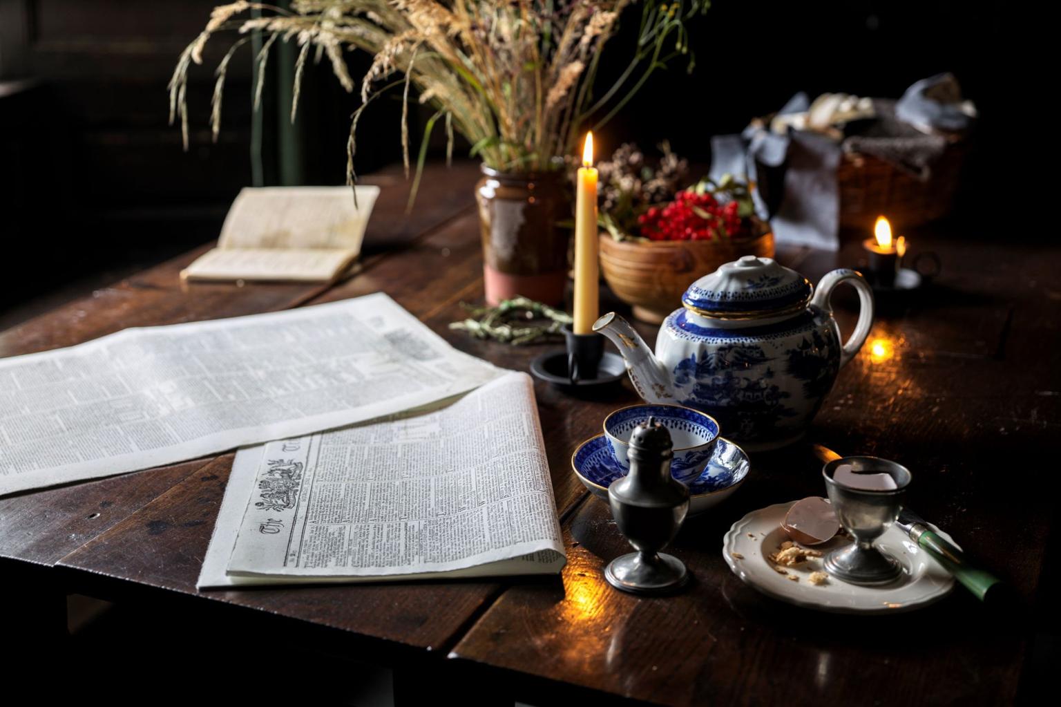 A teapot, teacup and saucer, egg cup, candles, newspapers and flowers on a dark wooden table.