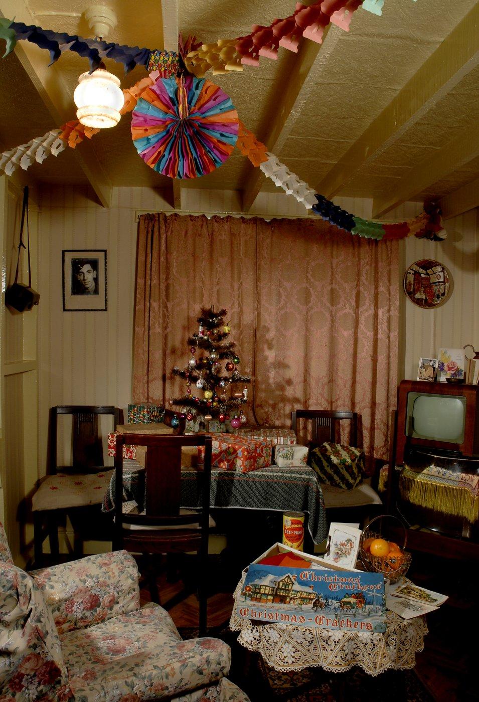 Period living room decorated for Christmas with a small lit tree on a table and multicolored paper garlands and fan hanging from the ceiling.