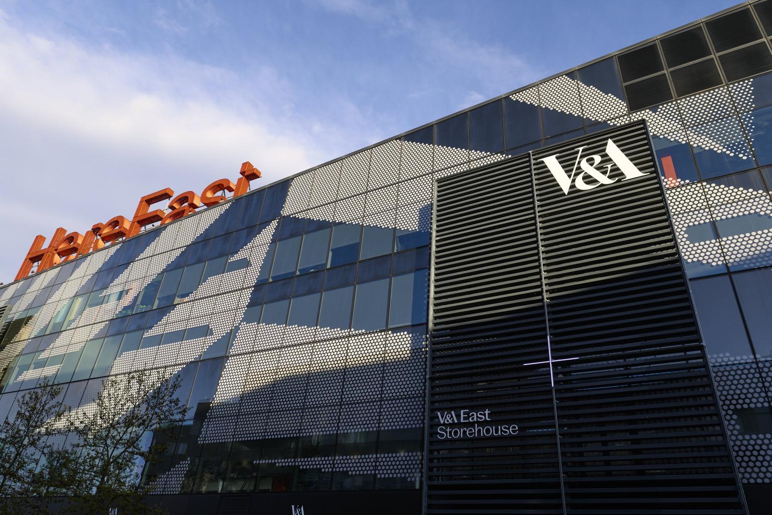The industrial glass and steel facade of the V&A East Storehouse with orange 'Here East' signage atop the building.