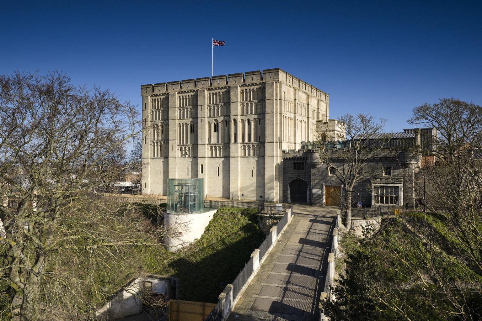 A pale stone medieval keep flying a Union Jack stands under a clear blue sky.