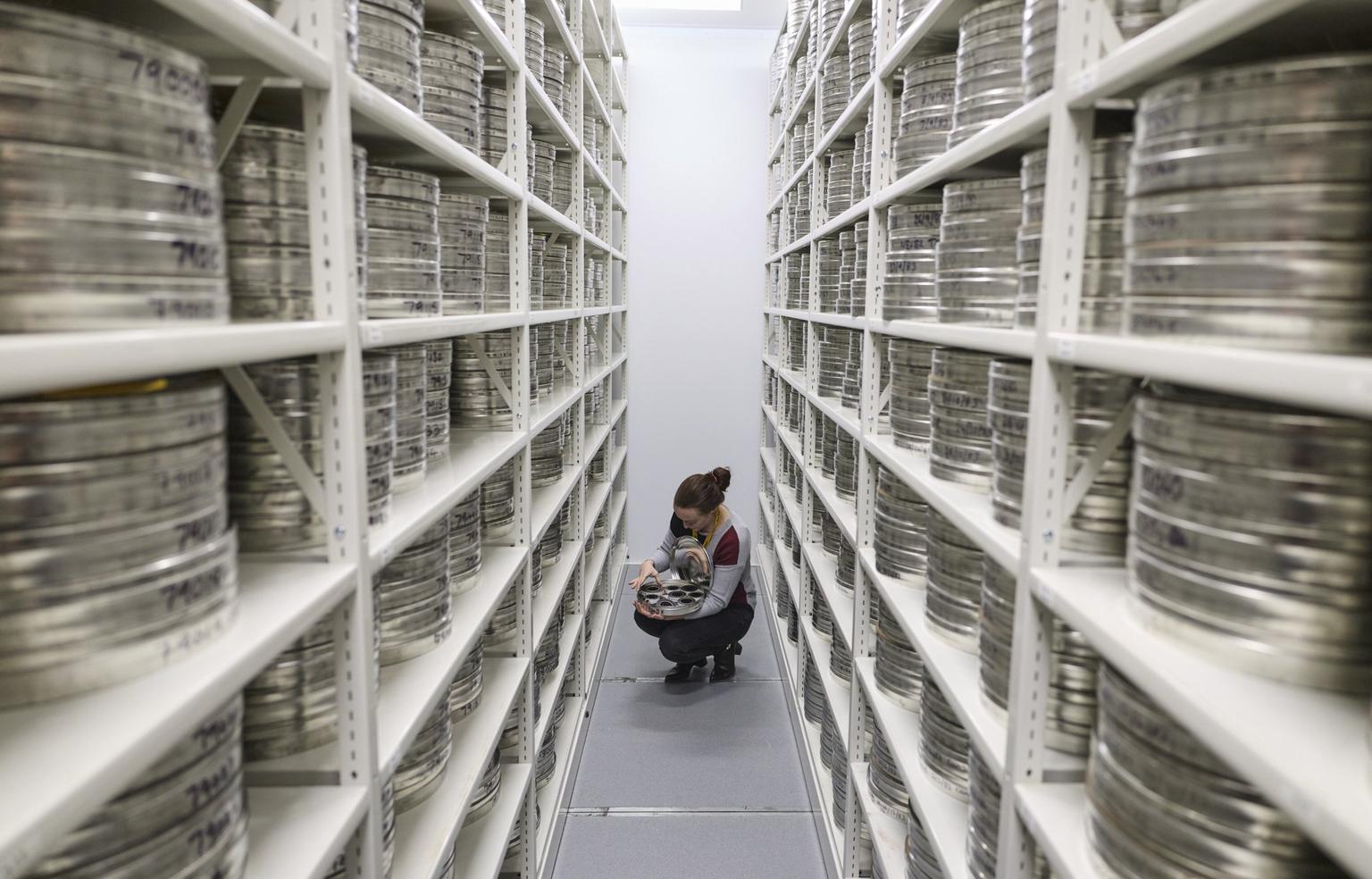 A person crouches between white shelving aisles inspecting stacks of silver film canisters in an archival storage room.